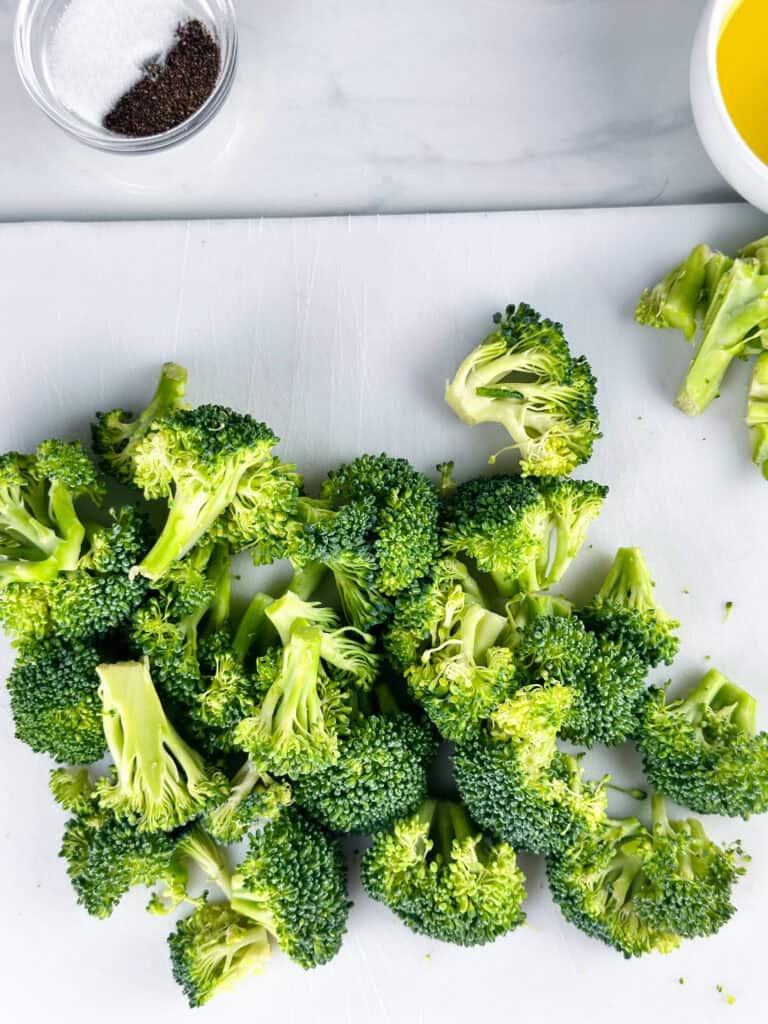 Broccoli on a white cutting board with olive oil and salt and pepper in background.