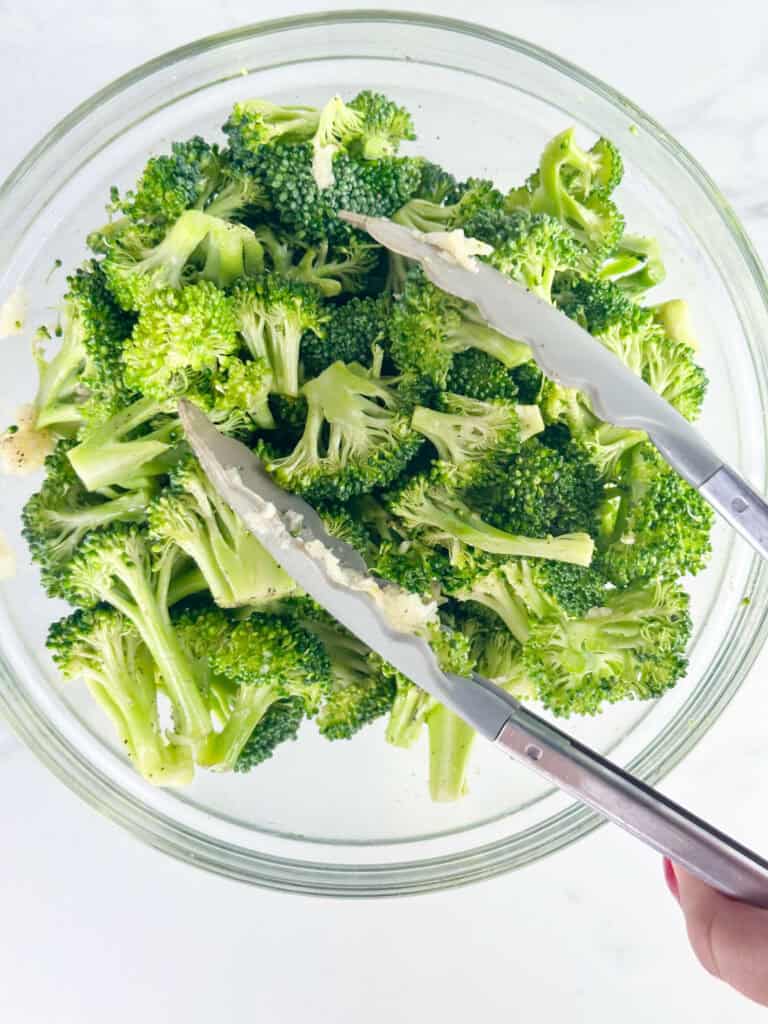 Tossing broccoli in a clear bowl with grey tongs on a white background.