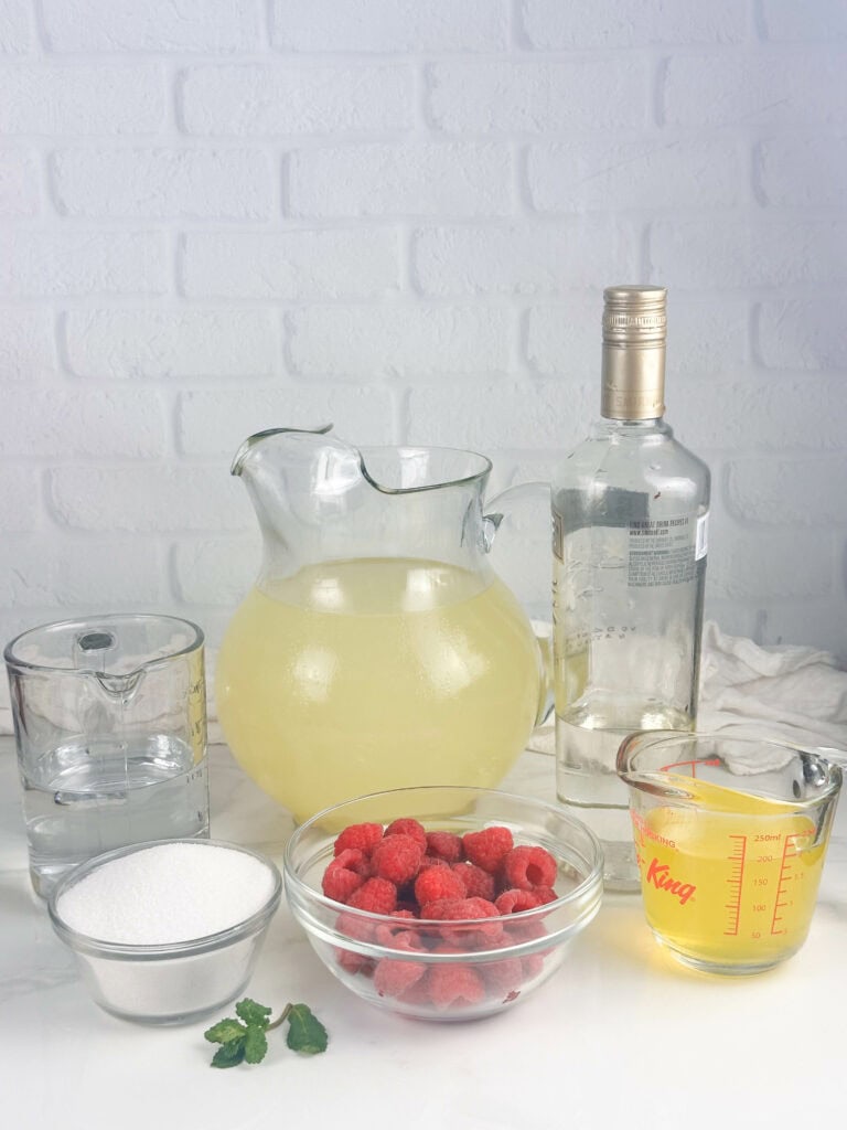 Raspberry lemonade cocktail ingredients on a white background. Pictured ingredients include: pitcher of lemonade, water, sugar, red raspberries, limoncello, vodka, and mint leaves.