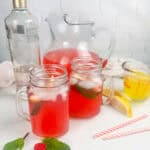Raspberry lemonade cocktail in pitcher and mason jars. Also pictured a bottle of vodka, straws, fresh raspberries, lemon wedges, and limoncello.