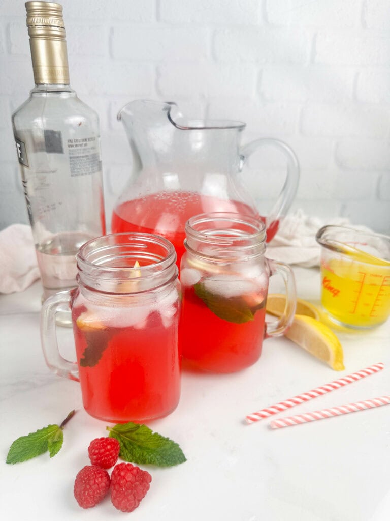 Raspberry lemonade cocktail in pitcher and mason jars. Also pictured a bottle of vodka, straws, fresh raspberries, lemon wedges, and limoncello.