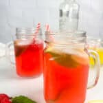 Two mason jars of raspberry lemonade cocktail on a white background with raspberries, mint leaves, lemon wedges, limoncello, and a bottle of vodka in the background.