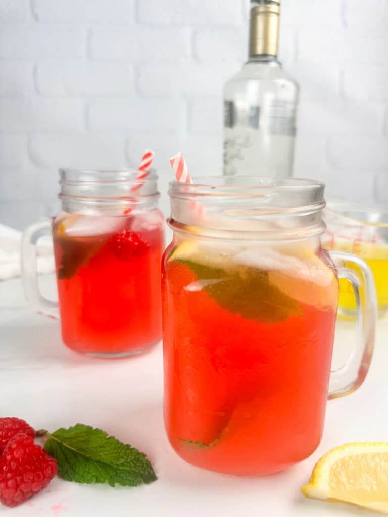 Two mason jars of raspberry lemonade cocktail on a white background with raspberries, mint leaves, lemon wedges, limoncello, and a bottle of vodka in the background.