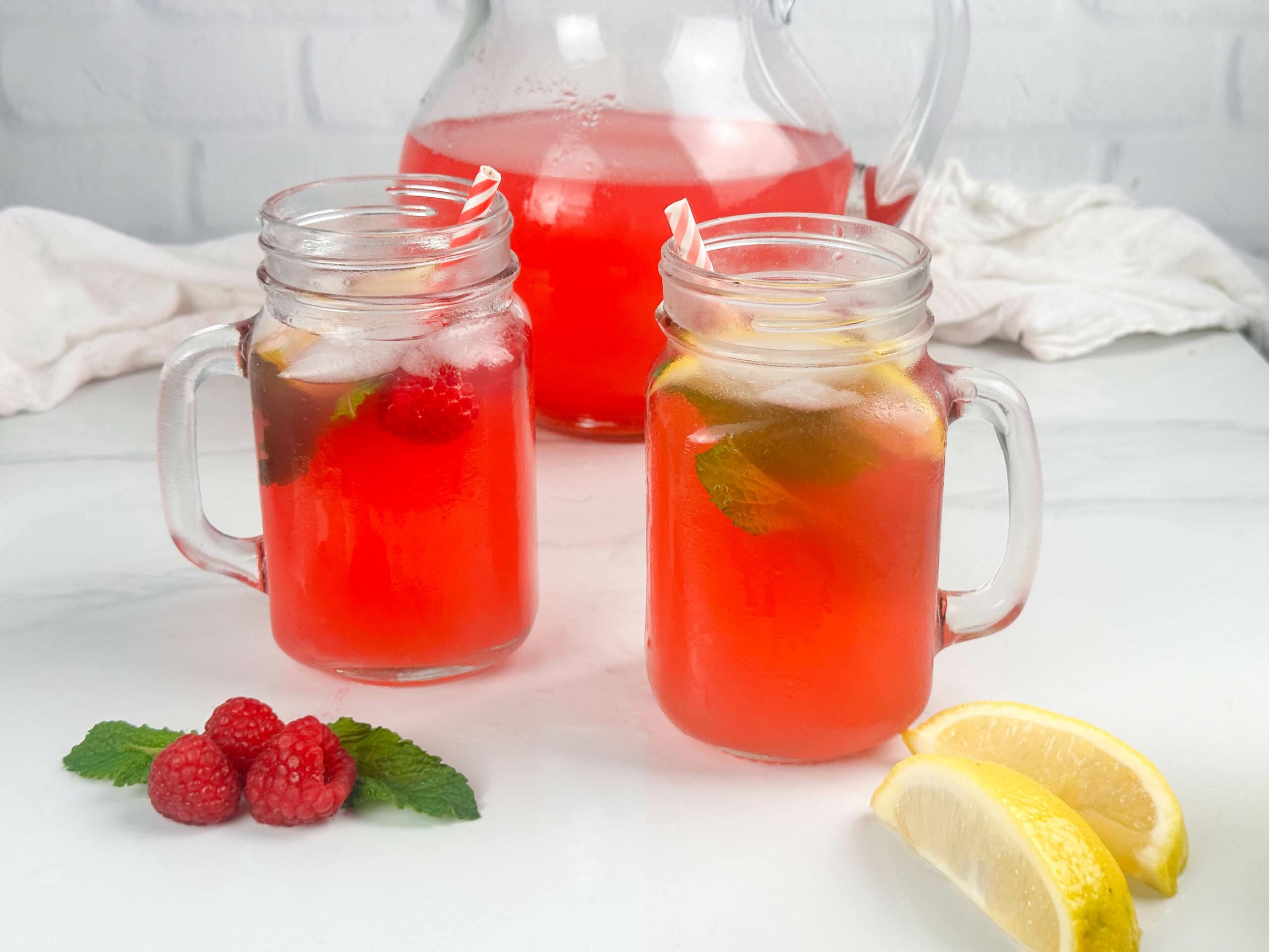 Two mason jars of raspberry lemonade cocktail on a white background with raspberries, mint leaves, lemon wedges, limoncello, and a bottle of vodka in the background.
