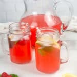 Red raspberry lemonade in two mason jar glasses with pitcher in the background and fresh raspberries, mint leaves, red and white straws, and lemon wedges in the foreground.