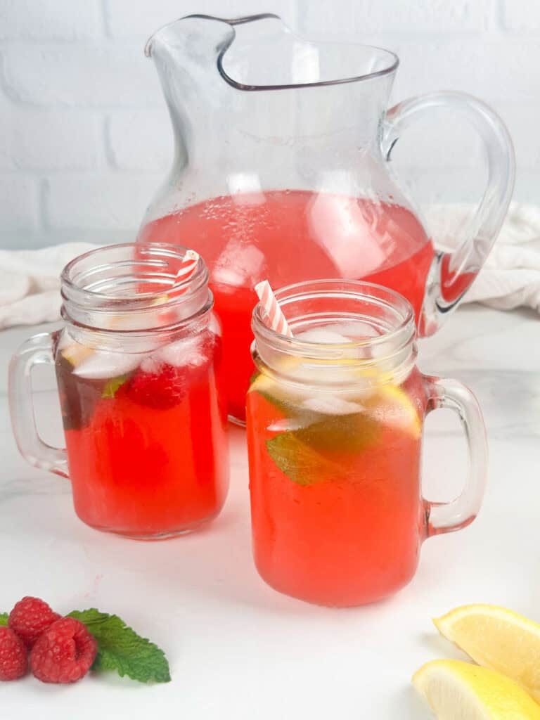 Red raspberry lemonade in two mason jar glasses with pitcher in the background and fresh raspberries, mint leaves, red and white straws, and lemon wedges in the foreground.