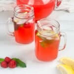 Red raspberry lemonade in two mason jar glasses with pitcher in the background and fresh raspberries, mint leaves, and lemon wedges in the foreground.