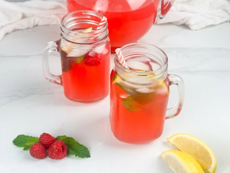 Red raspberry lemonade in two mason jar glasses with pitcher in the background and fresh raspberries, mint leaves, and lemon wedges in the foreground.