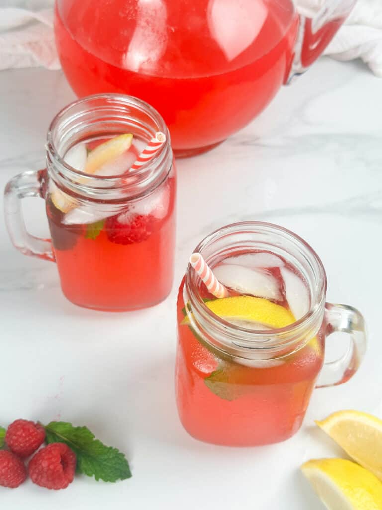 Red raspberry lemonade in two mason jar glasses with pitcher in the background and fresh raspberries, mint leaves, and lemon wedges in the foreground.