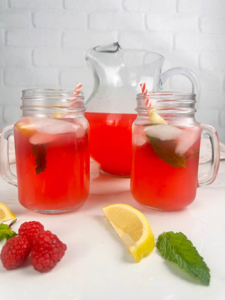 Red raspberry lemonade in two mason jar glasses with pitcher in the background and fresh raspberries, mint leaves, and lemon wedges in the foreground.