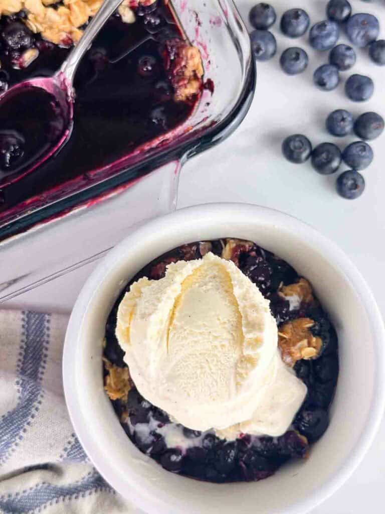 Scoop of vanilla ice cream on top of blueberry crumble in a white bowl.