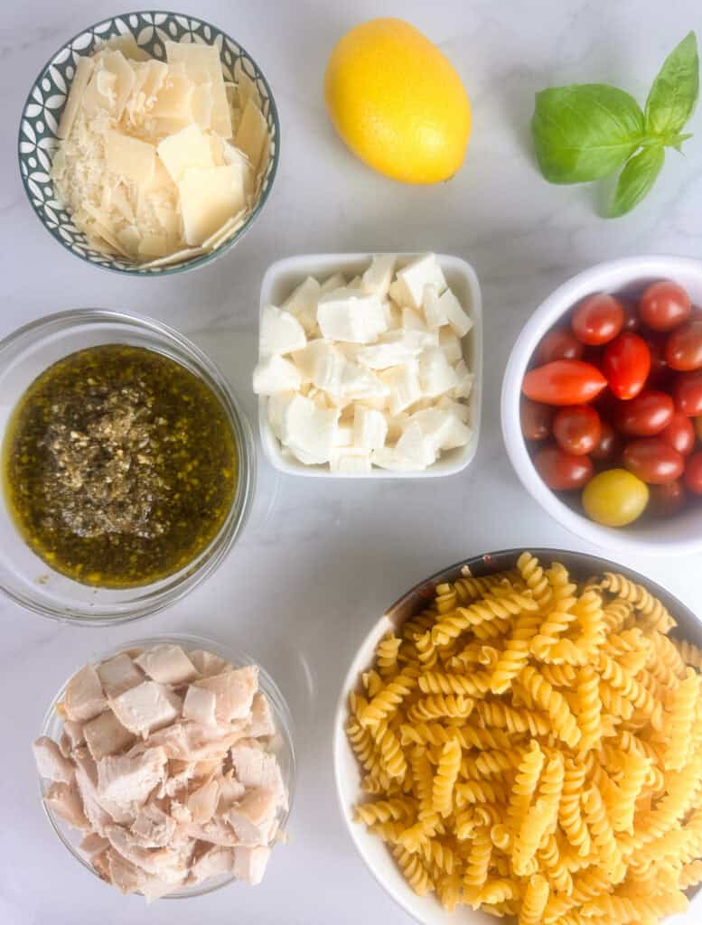 Ingredients for chicken pesto pasta salad: parmesan cheese, lemon, mozzarella cheese, pesto sauce, cherry tomatoes, chicken, and pasta.