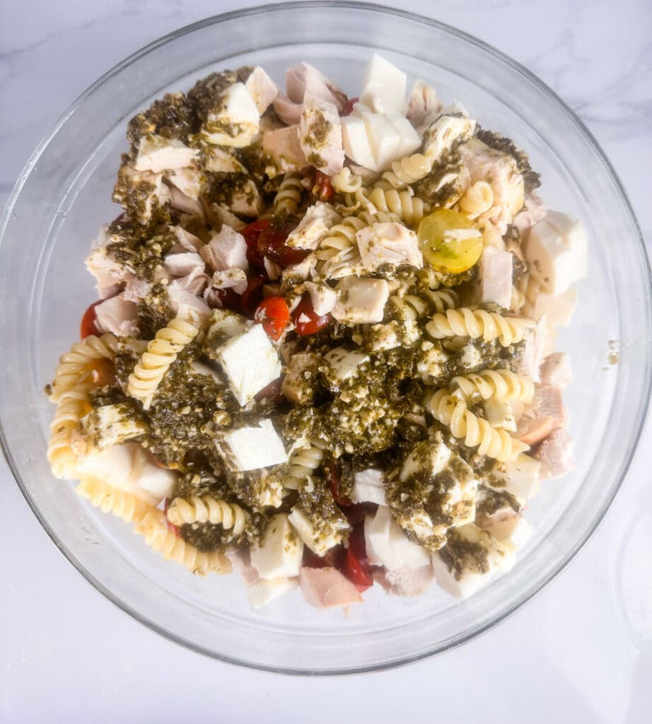 Ingredients for chicken pesto pasta salad added to a clear bowl, ready to mix.