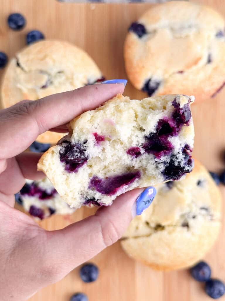 Hand holding half of a buttermilk blueberry muffin over other muffins on a wood cutting board.