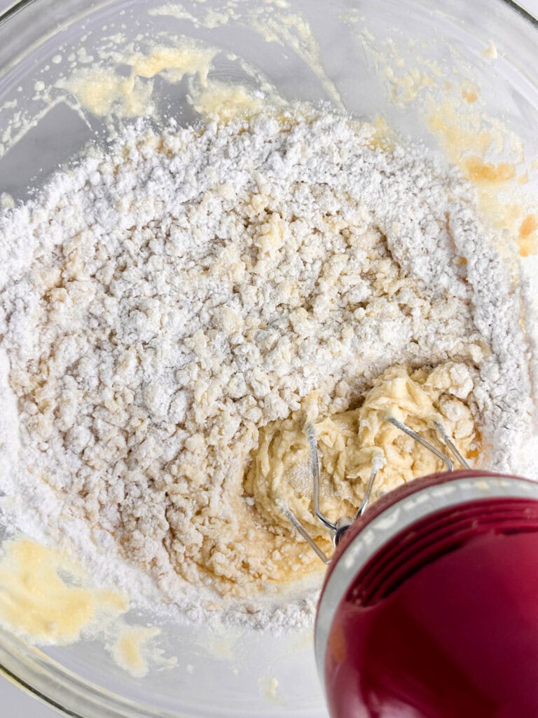 Combining flour with creamed butter in a clear mixing bowl with a red hand mixer.