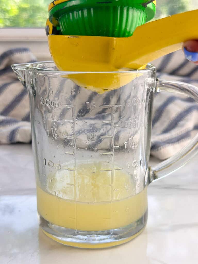 Handheld lemon juicer squeezing a lemon into a clear measuring cup in front of a window and towel.