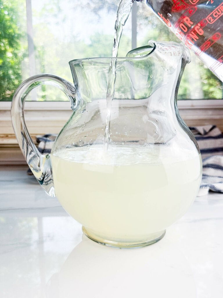 Clear pitcher with lemon juice and simple syrup getting filled with water in front of a window.