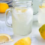 Mason jar mug of homemade lemonade with lemon wedges, a lemon, and a pitcher in the background.