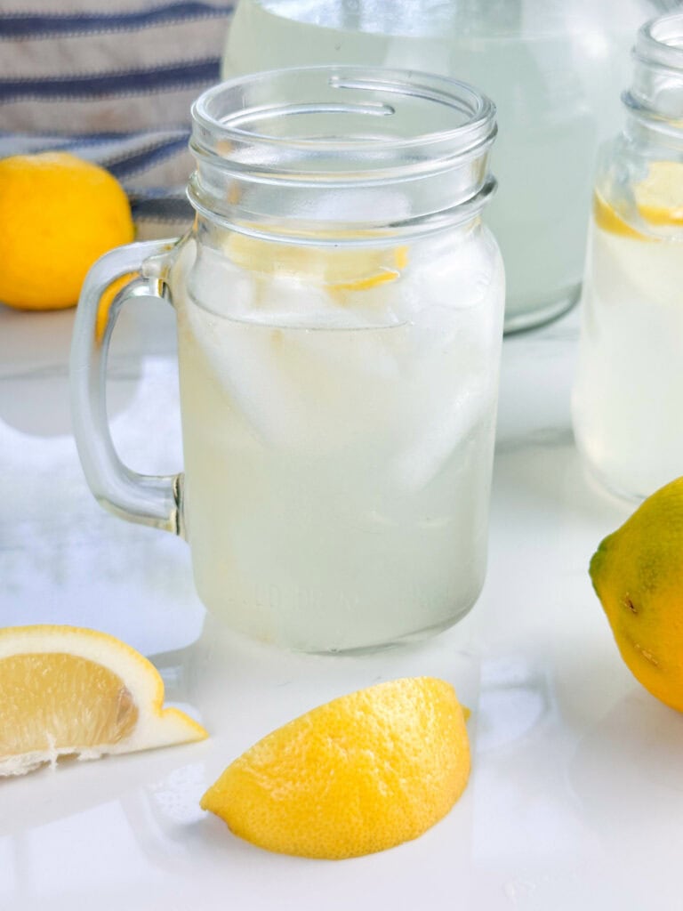 Mason jar mug of homemade lemonade with lemon wedges, a lemon, and a pitcher in the background.