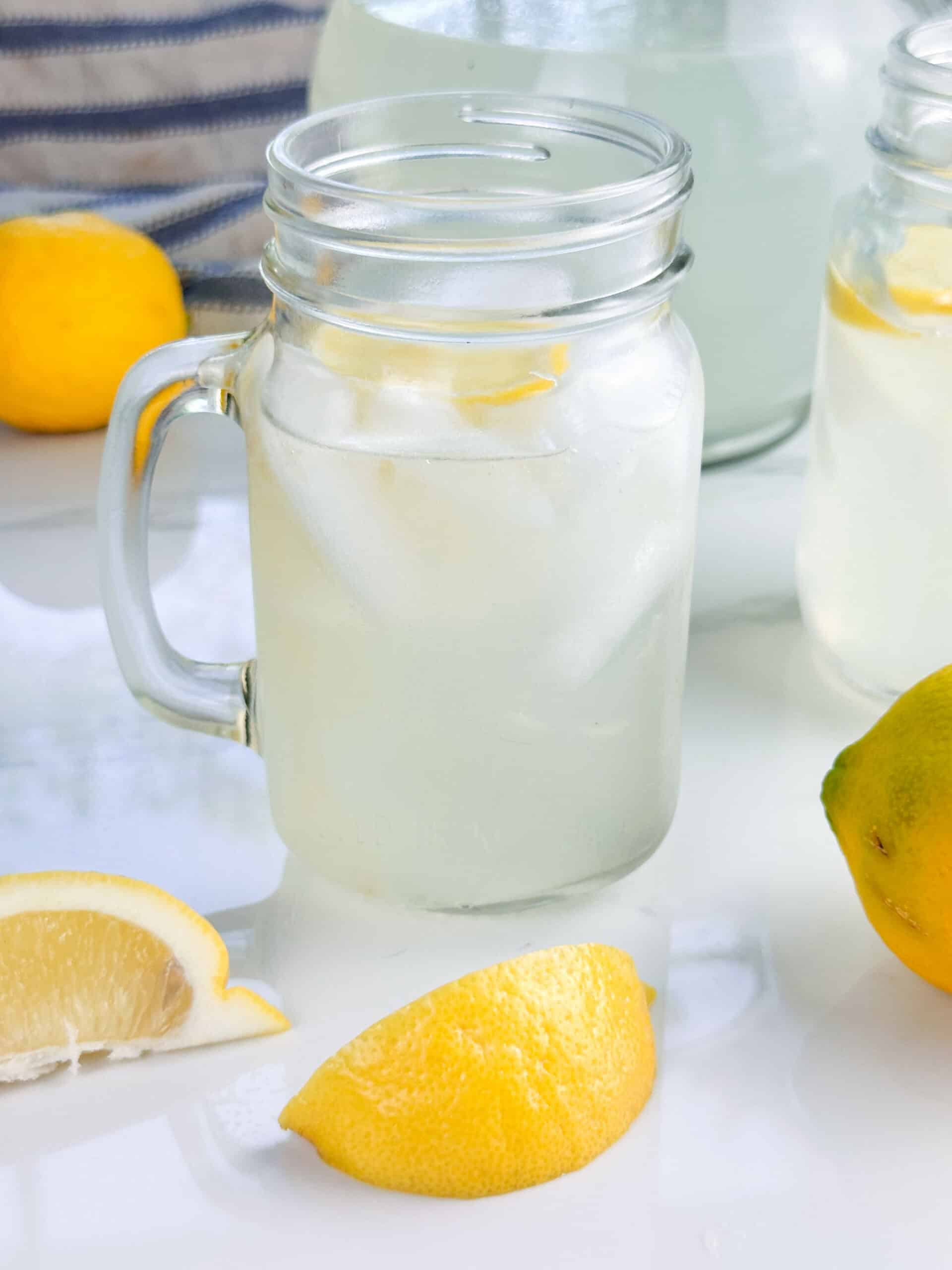 Mason jar mug of homemade lemonade with lemon wedges, a lemon, and a pitcher in the background.