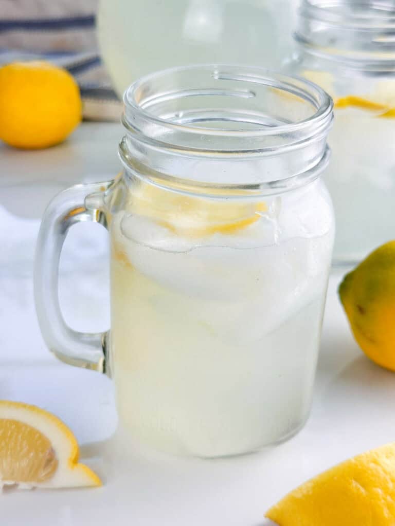 Single mason jar mug of homemade lemonade with lemons, an additional glass, and pitcher in the background.