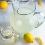 Pitcher and mason jar cup of lemonade on a white background with lemons in the background.