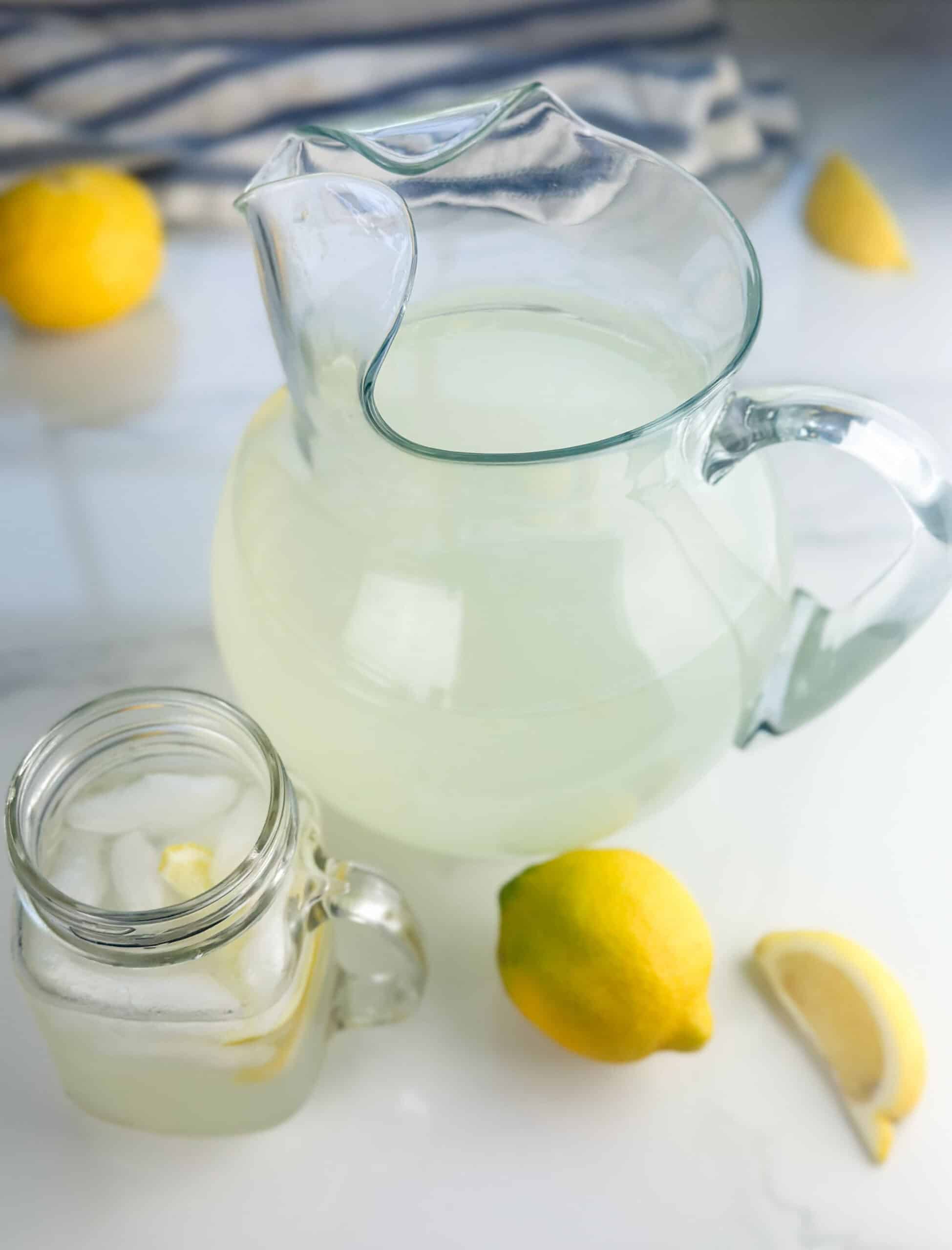 Pitcher and mason jar cup of lemonade on a white background with lemons in the background.