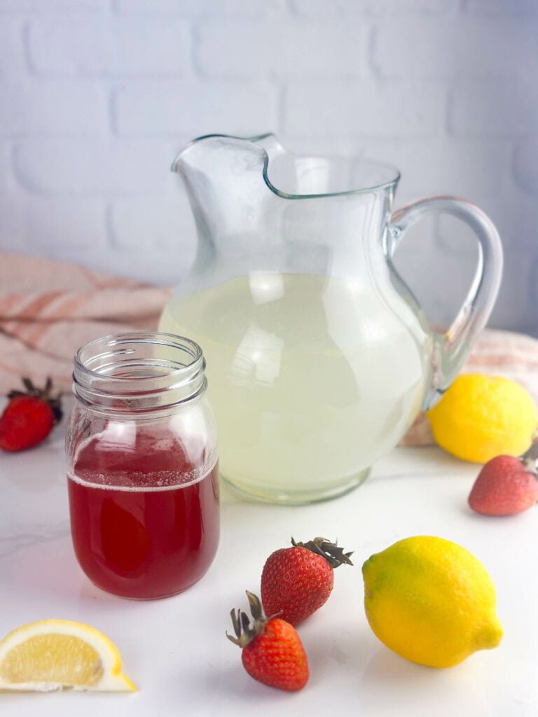 Ingredients for a homemade strawberry lemonade recipe: lemonade and strawberry simple syrup. Lemons and strawberries fill out the shot on a white background.