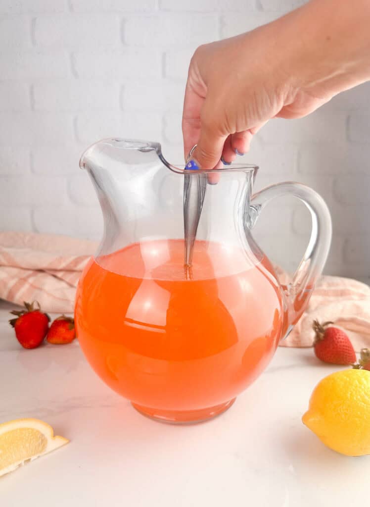 Hand stirring strawberry lemondade with a metal spoon on white background.