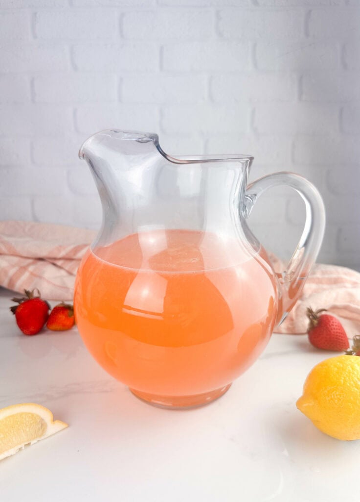 Strawberry lemonade in a clear pitcher with lemons and strawberries set on a neutral background.