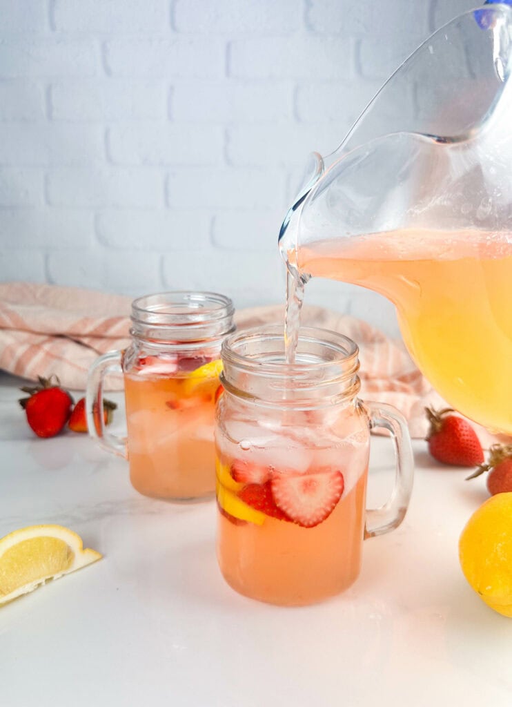 Pouring strawberry lemonade into waiting mason jar glasses with ice, strawberry, and lemon wedges.