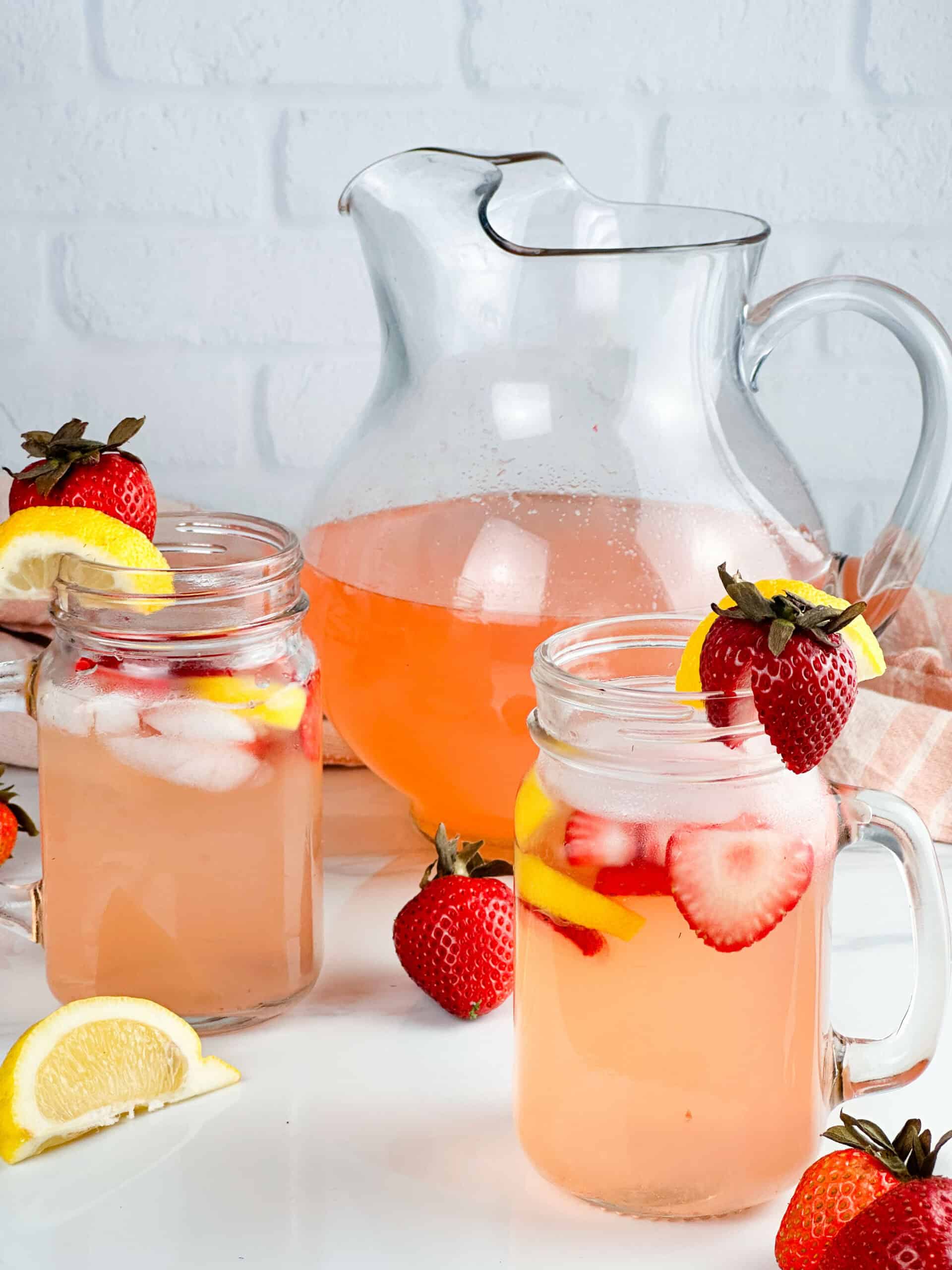 Two glasses and a pitcher of strawberry lemonade on a neutral background.
