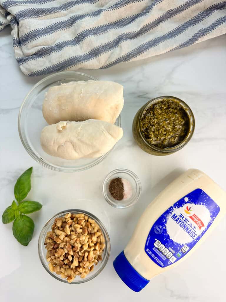 Pesto chicken salad ingredients on a white tile surface: cooked chicken, pesto sauce, basil leaves, walnuts, jar of mayonnaise, and salt and pepper.