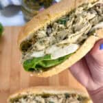 Hand holding pesto chicken salad sandwich over a cutting board with another sandwich on it.
