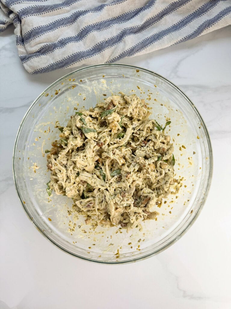 Shredded chicken mixed with mayo, pesto, basil leaves, and walnuts in a clear bowl on a white tile.