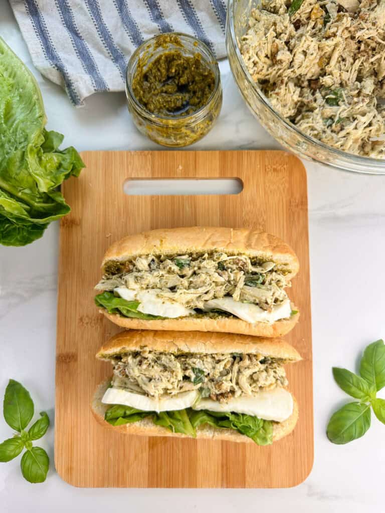 Two pesto chicken salad sandwiches on a wooden cutting board with a jar of pesto sauce and bowl of chicken salad to the side. Lettuce and basil also shown.