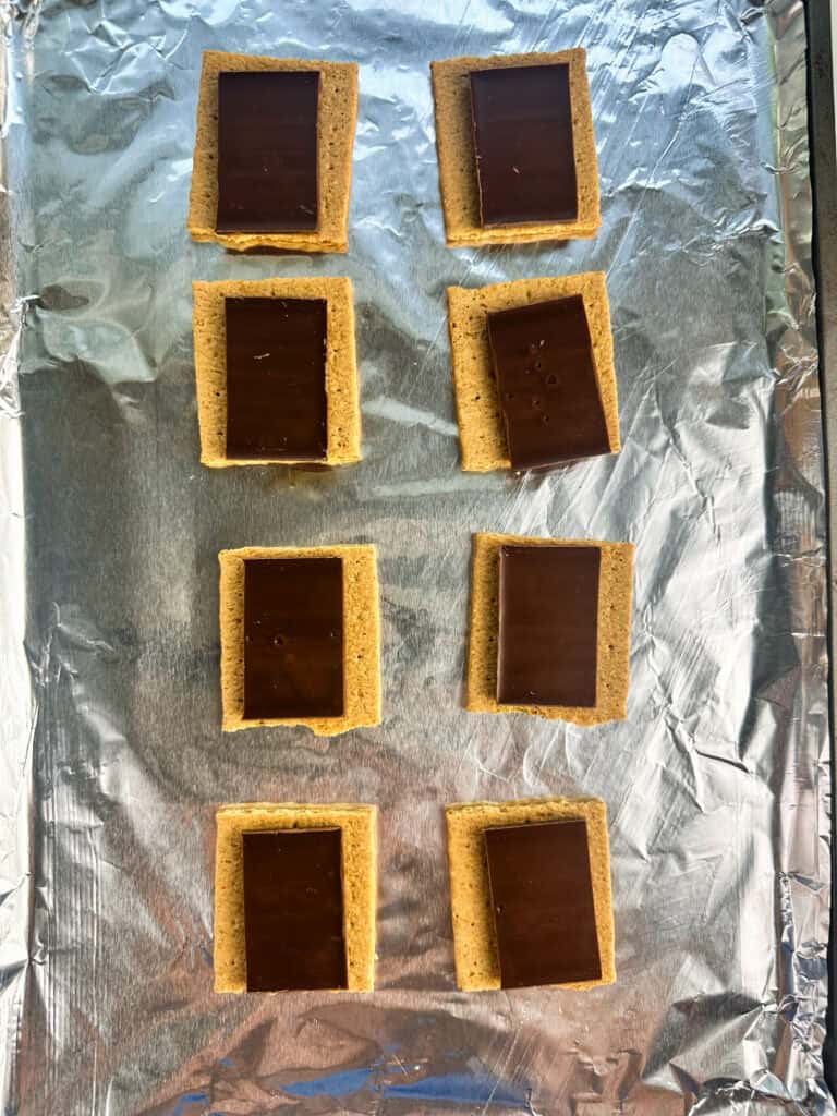 Graham cracker squares arranged on a sheet pan lined with aluminum foil with a square of chocolate bar on top.