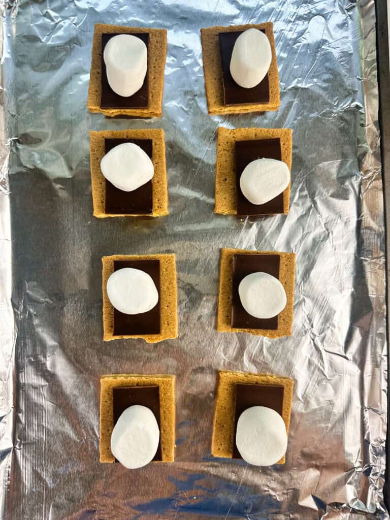 Graham cracker squares arranged on a sheet pan lined with aluminum foil with a square of chocolate bar and marshmallow on top.