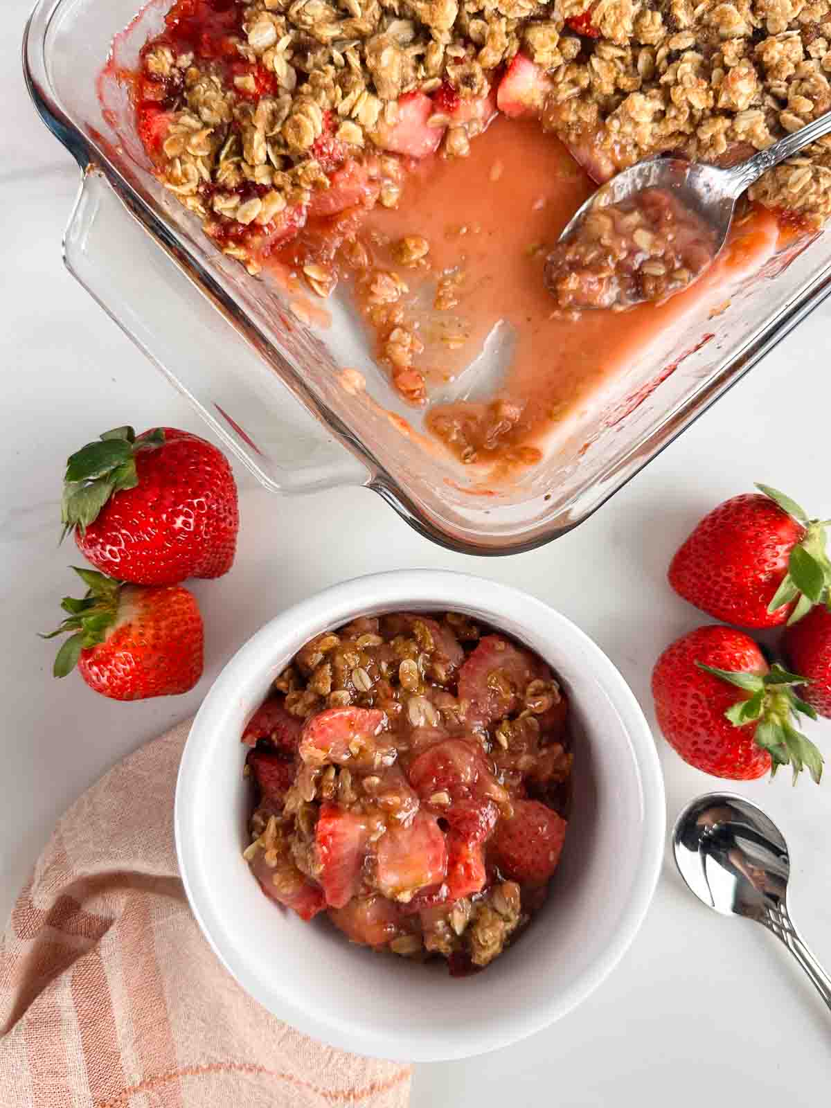Serving of strawberry crumble in a white bowl with the baking dish of strawberry crumble in background.