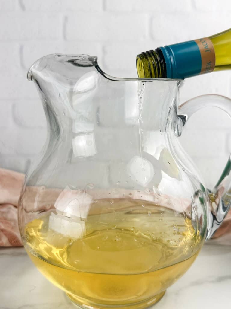 Pouring white wine into a clear pitcher in front of a neutral background.