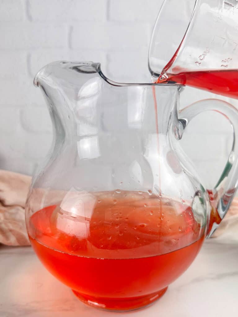 Pouring strawberry simple syrup into a clear pitcher in front of a neutral background.