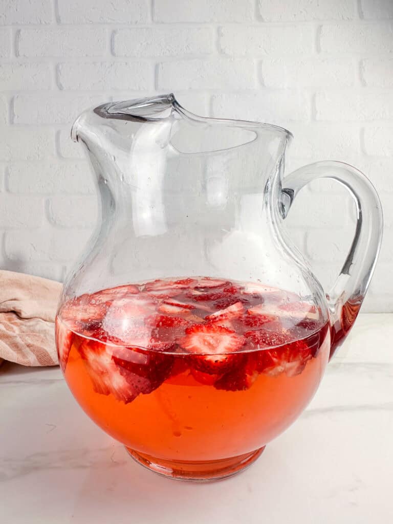 Sliced strawberries in a clear pitcher in front of a neutral background.