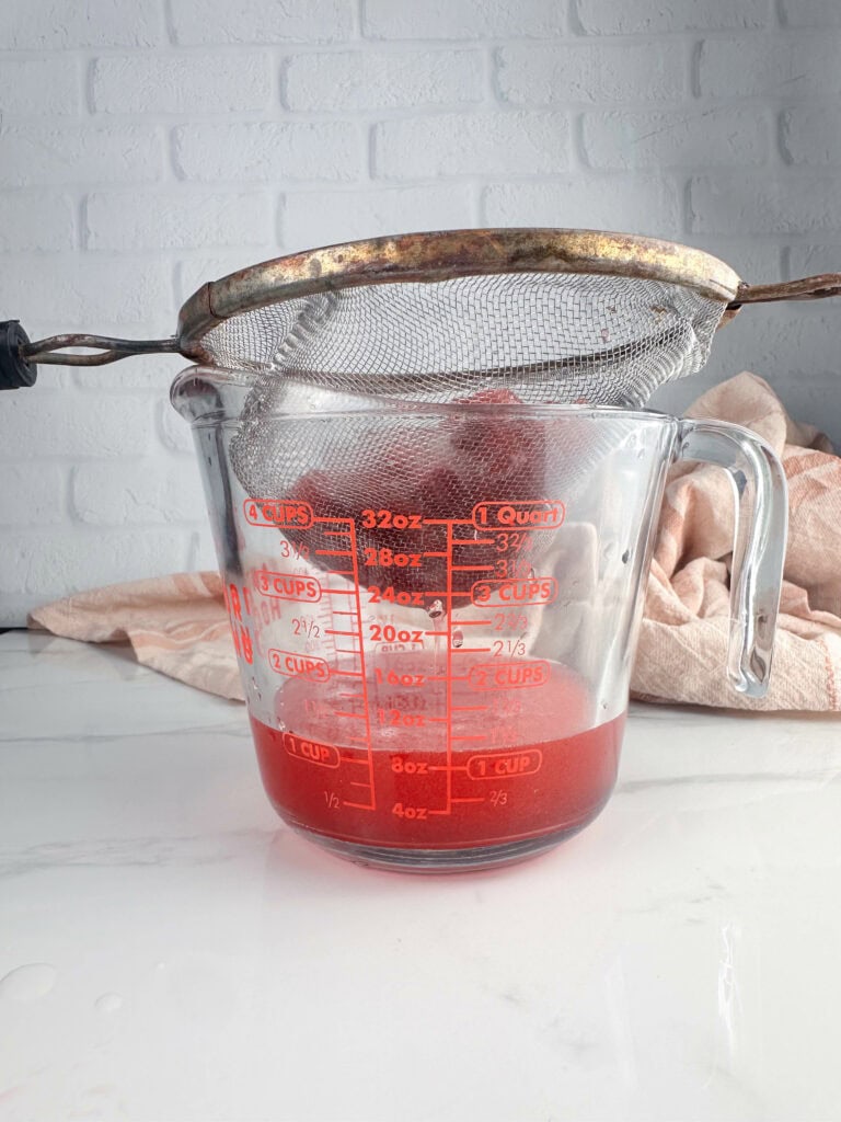 A mesh strainer set over a measuring cup to strain the solids out of the strawberry simple syrup