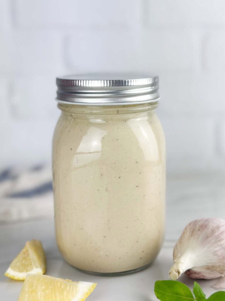 Jar of Caesar salad dressing with lemon, garlic, and basil on a neutral background in a sealed mason jar.