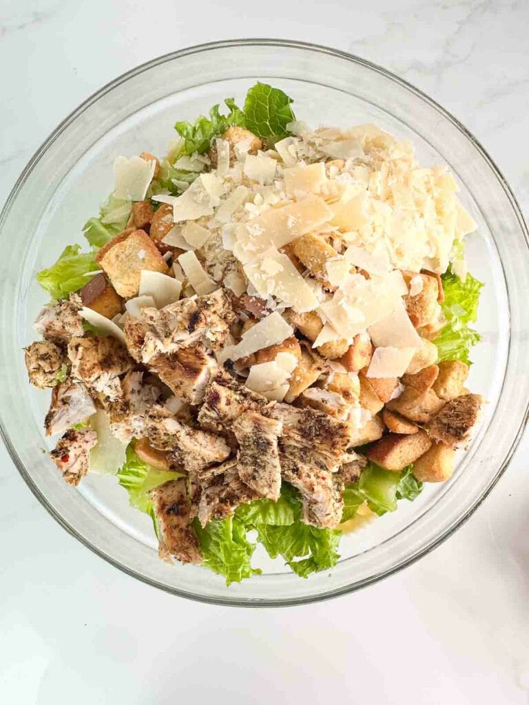 Croutons, lettuce, chicken, parmesan cheese, and noodles added to a clear mixing bowl.