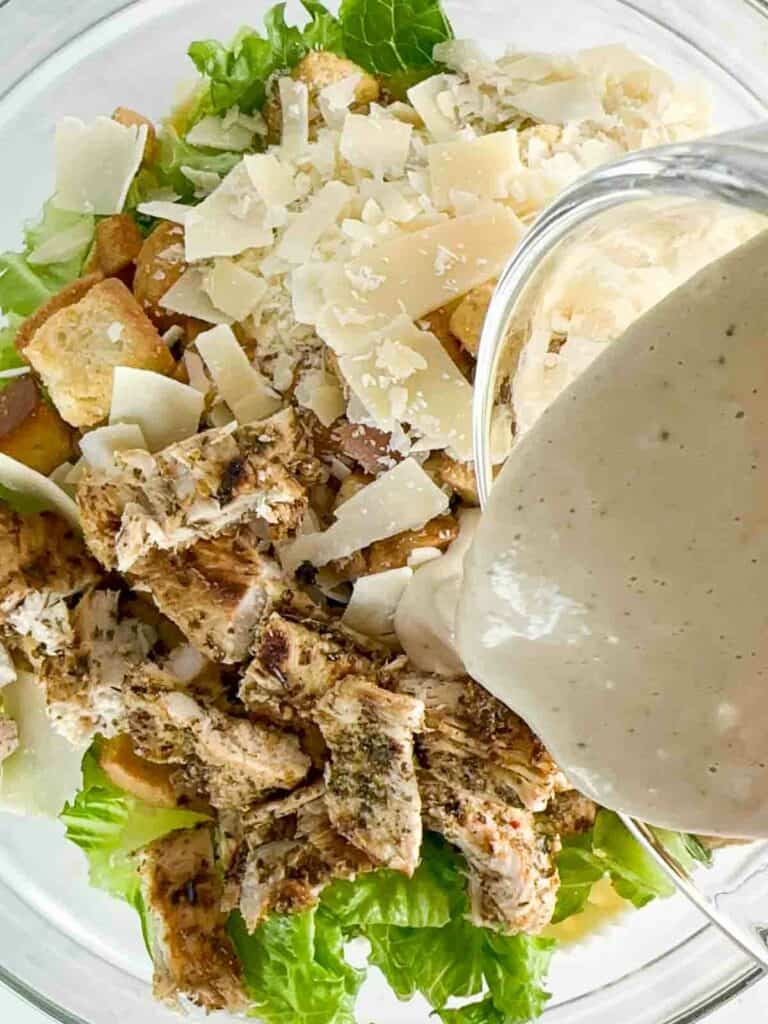 Pouring Caesar dressing over chicken, croutons, cheese, lettuce, and noodles for pasta salad.