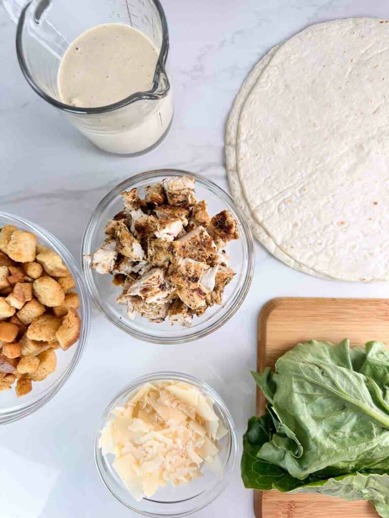 Ingredients for chicken Caesar salad wrap: Caesar dressing, flour tortillas, grilled or cooked chicken, croutons, shaved parmesan cheese, and romaine lettuce.