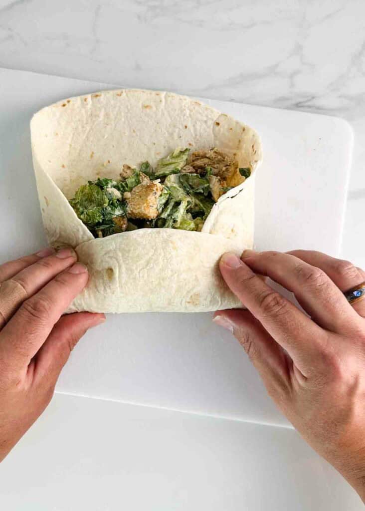 Folding a chicken caesar salad wrap on a white cutting board.