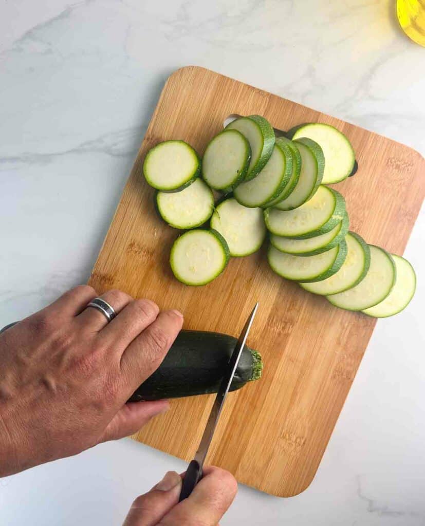 Slicing the ends off the zucchini with a sharp knife on a wood cutting board with zucchini rounds.