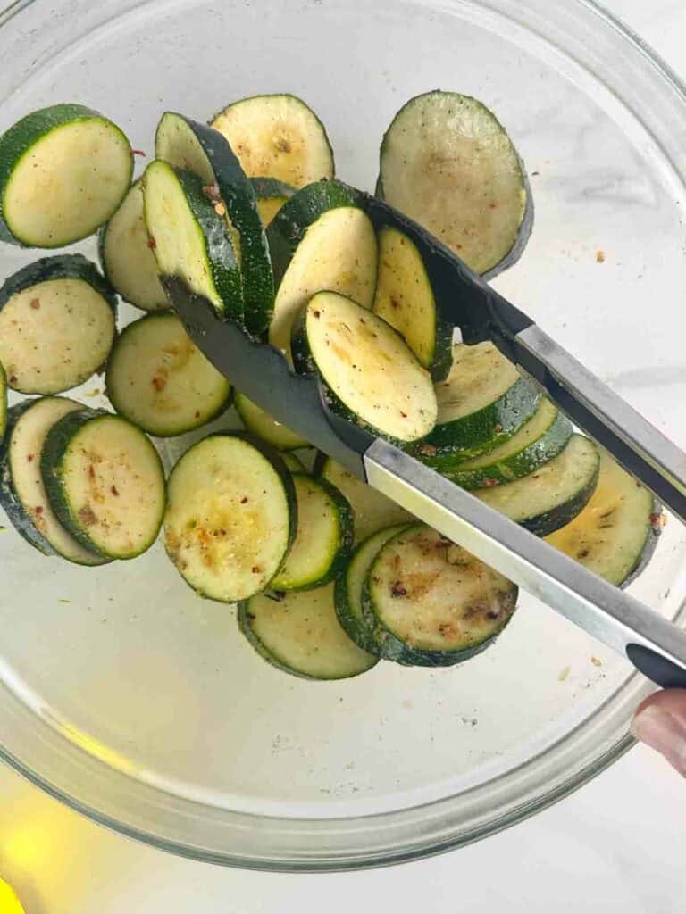 Mixing zucchini rounds with tongs in a clear bowl.